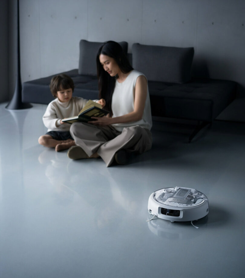 A woman and child sit on the floor reading a book while a DJI Romo robotic vacuum cleaner operates nearby in a modern living room.