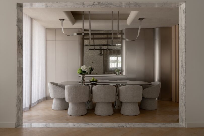 Modern dining room with a marble-framed entry, oval table, eight upholstered chairs, minimalist decor, large mirror, and neutral color tones.