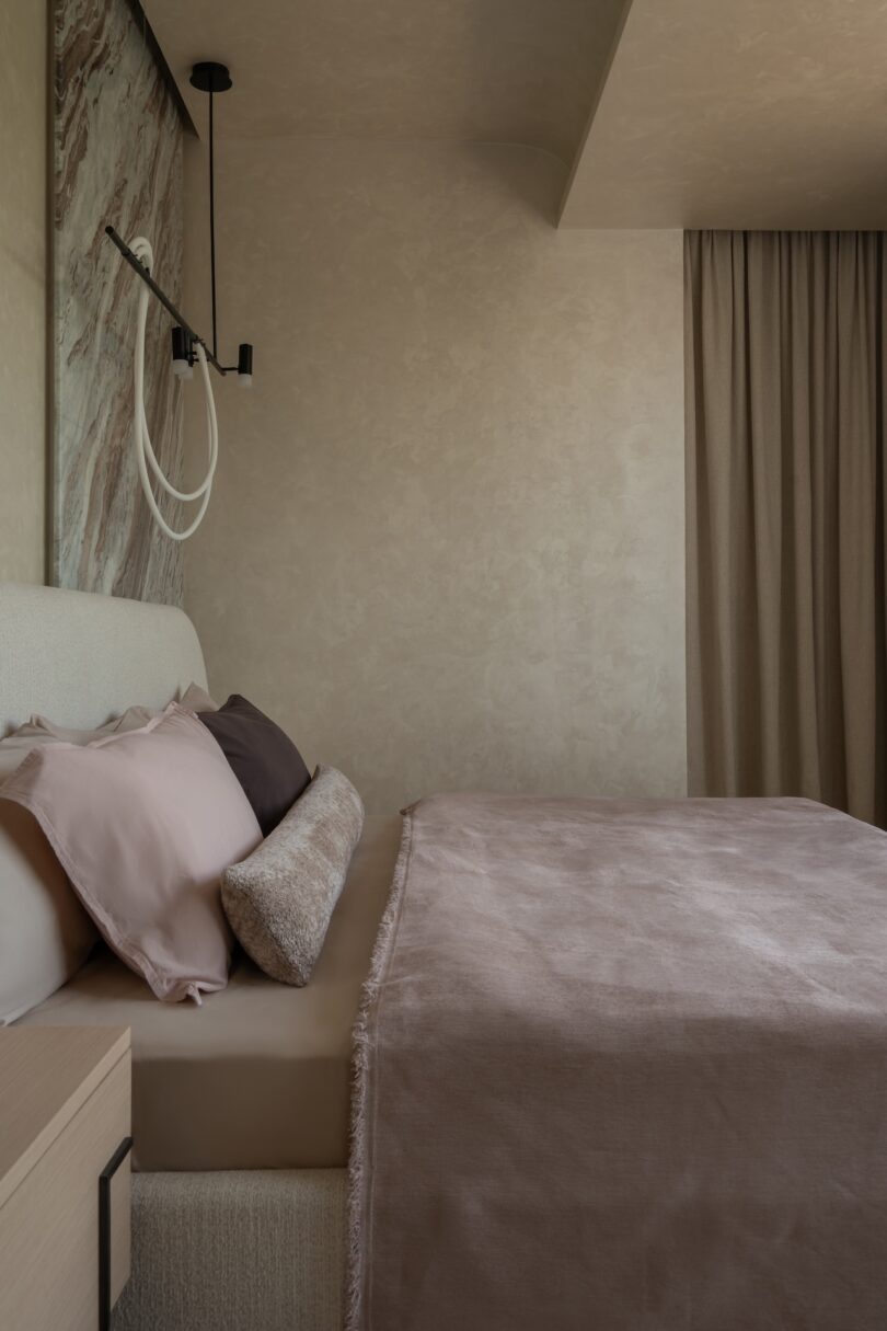 A minimalist bedroom with neutral tones, featuring a made bed with pink and brown bedding, a modern wall light, and floor-length curtains.