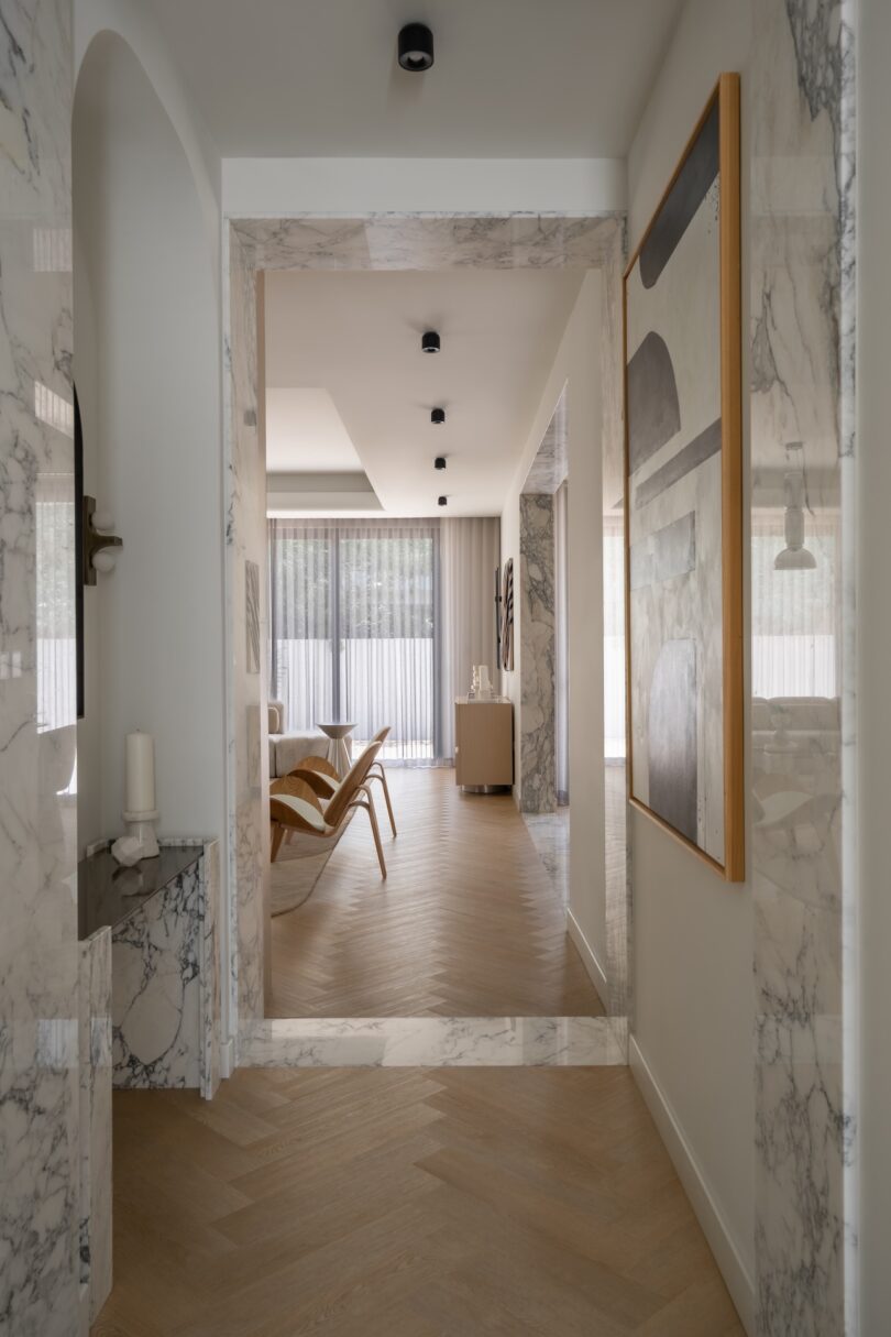 A hallway with marble walls leads to a bright living room with wooden floors, modern furniture, large abstract art, and floor-to-ceiling windows covered by sheer curtains.
