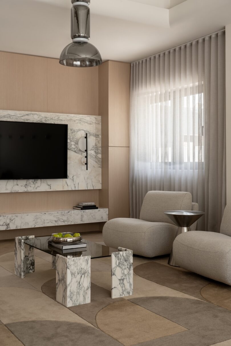 Modern living room with marble TV wall, marble coffee table, two beige armchairs, metallic side table, and large window with sheer curtains letting in natural light.