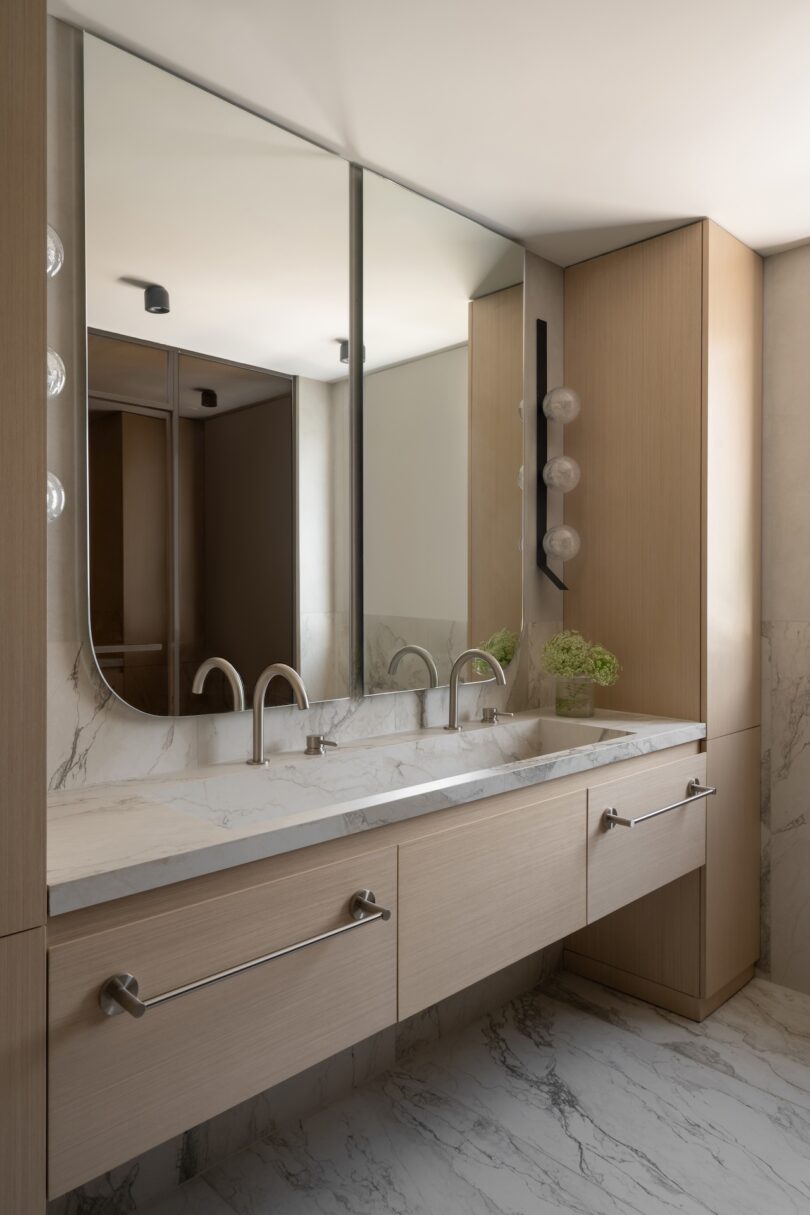 Modern bathroom with a double sink vanity, marble countertop and backsplash, large mirrors, wood cabinetry, and minimal lighting fixtures.
