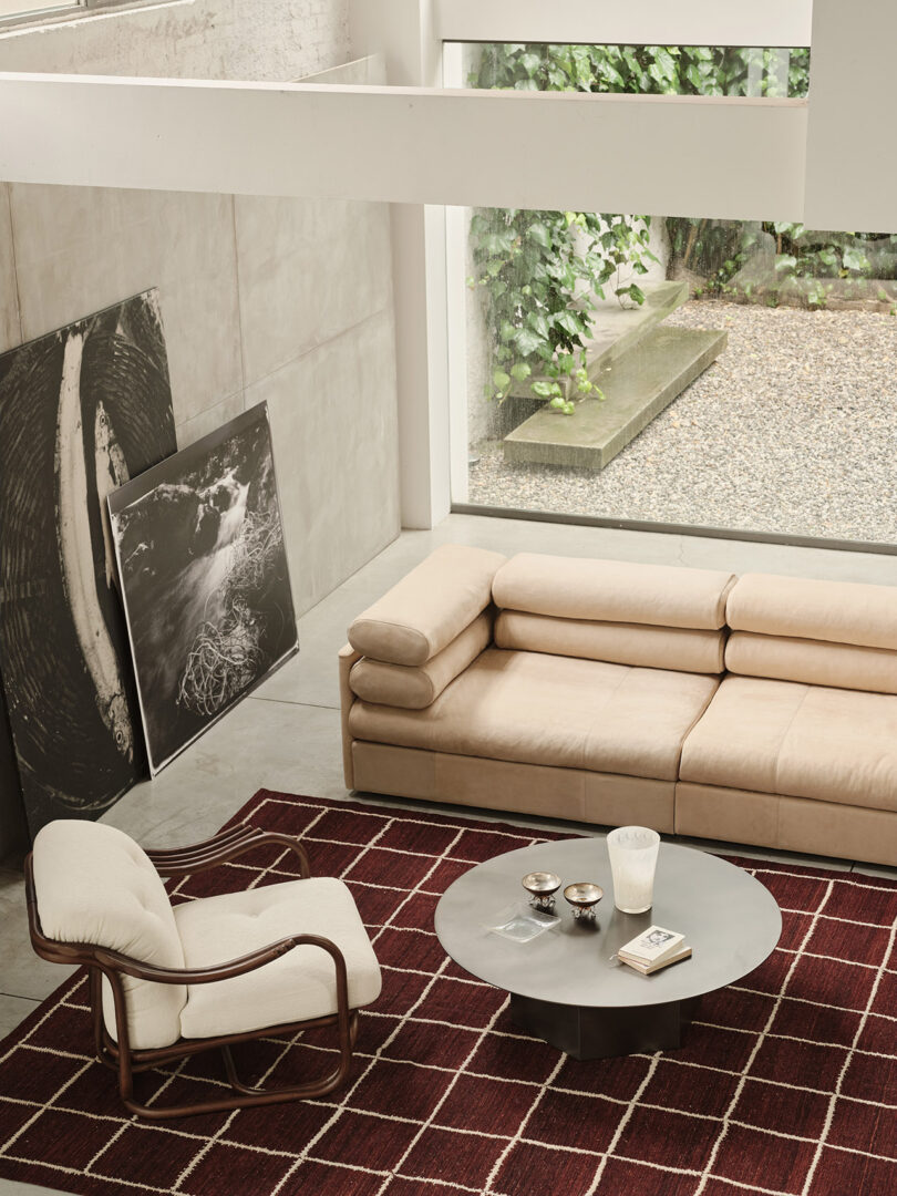 Modern living room with a beige sofa, cream armchair, round coffee table, and red geometric rug; large window shows greenery outside, and abstract art rests against a concrete wall.