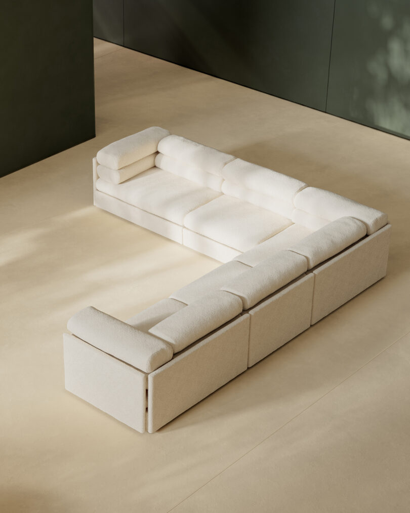 A modern beige sectional sofa with tufted cushions is arranged in an open, minimalist room with light flooring and dark green walls.