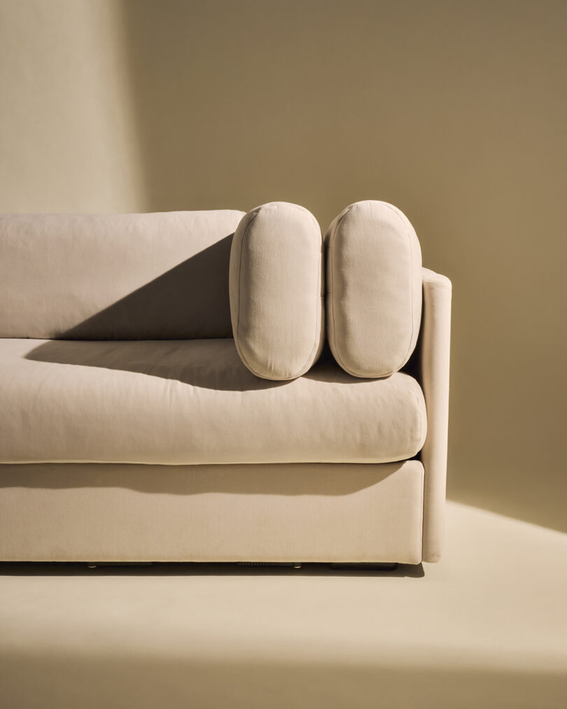 A beige upholstered sofa with two cylindrical cushions placed vertically on one armrest, set against a neutral background.