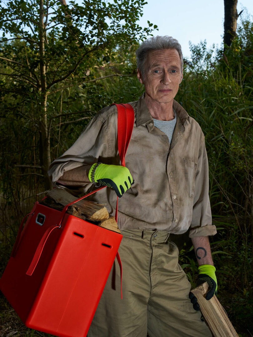 A man wearing a dirty shirt and green gloves carries firewood and a large red container in a wooded outdoor area.