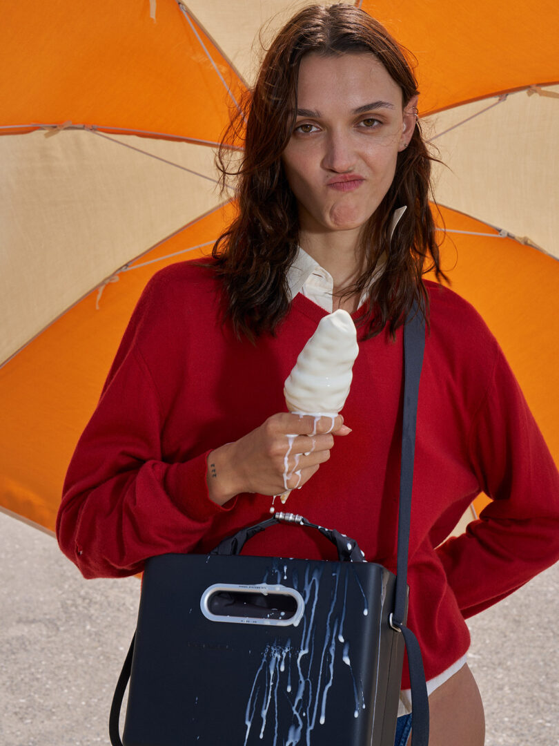 A woman in a red sweater holds a melting ice cream cone, with ice cream dripping onto her black bag, standing under a large orange umbrella.
