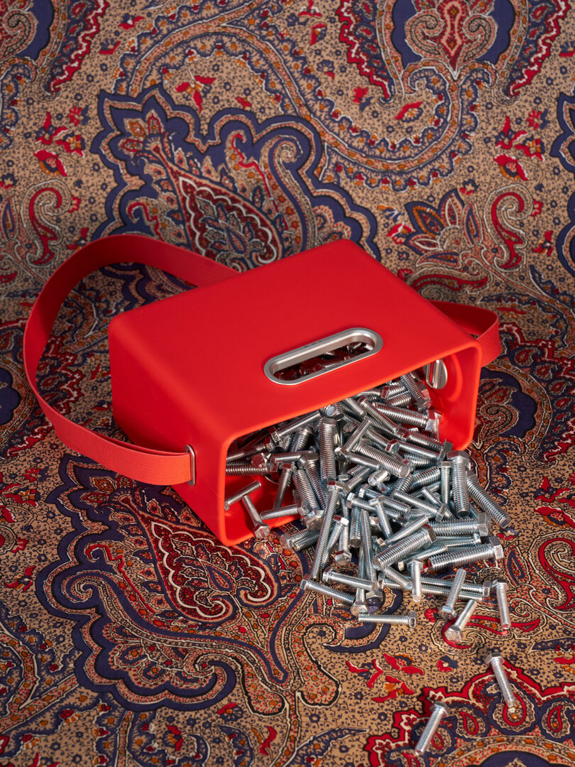 A red bag with a handle tipped over on a patterned surface, spilling metal bolts and screws onto the fabric.