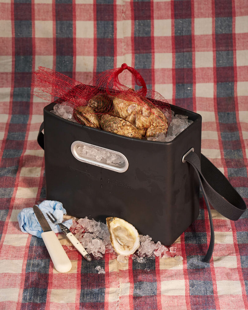 A black bag filled with ice and oysters sits on a red and white plaid cloth. Oyster knives, gloves, and opened oyster shells are scattered nearby.