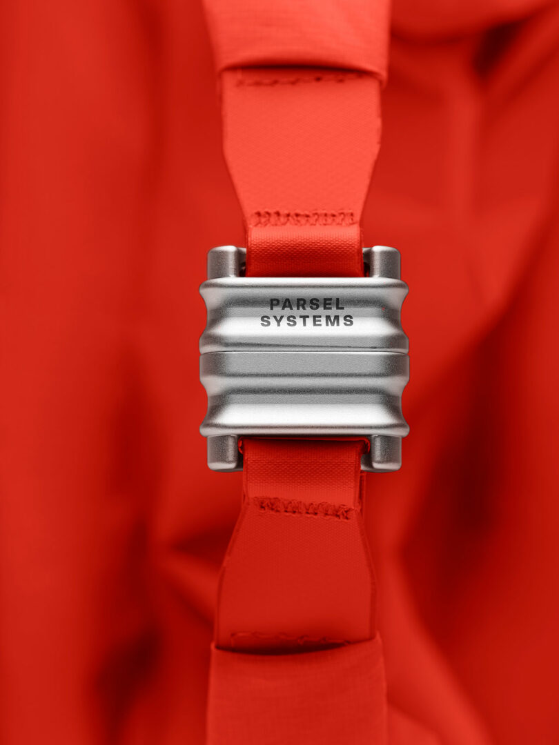 Close-up of a metallic buckle labeled "PARSEL SYSTEMS" attached to a red strap, part of the PARSEL EVA Tote System, set against a matching red background.