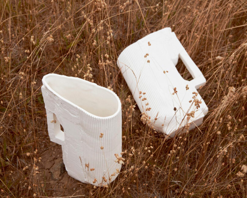 Two large, white, ceramic Dana Harel mugs with textured surfaces and square handles rest on dry grass among tall, brown plants.