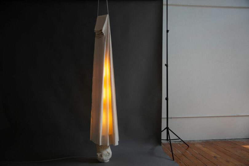A tall, cone-shaped Dana Harel lamp with folded fabric emits warm light, standing on a small white base against a dark backdrop in a studio setting.