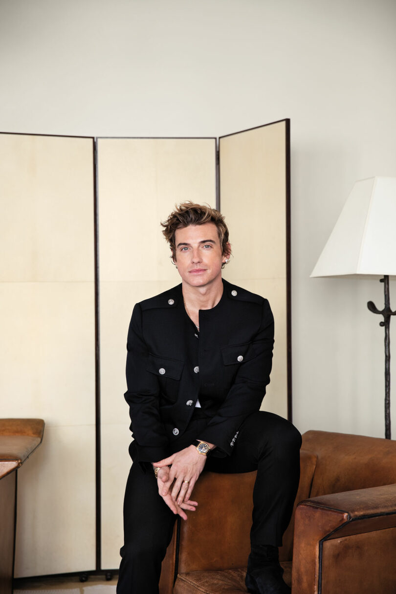 A person in a black buttoned jacket sits on a brown leather chair, with a cream-colored folding screen and a floor lamp in the background.