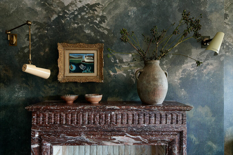 A Jeremiah Brent-inspired marbled fireplace mantel with two ceramic bowls and a large vase with twigs, beneath a framed painting and flanked by two wall-mounted lamps against a green patterned wall.