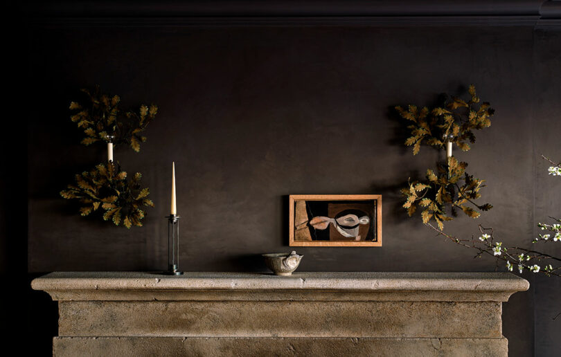 A stone mantel with a bowl, a glass candlestick with a white candle, wall sconces with gold leaves, and a framed abstract painting on a dark wall—an elegant look inspired by Jeremiah Brent.