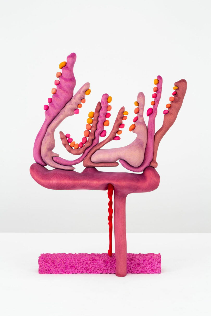 Abstract pink and purple sculpture by Laura Casañas Maya, featuring branching forms and small orange and pink spheres, stands on a textured pink base against a plain white background.