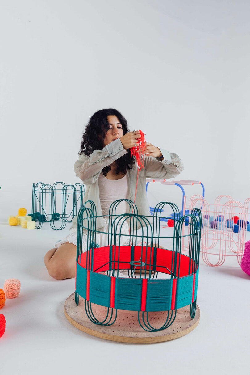 Laura Casañas Maya sits on the floor, weaving red yarn onto a circular metal frame; other colorful yarn and similar structures surround her.