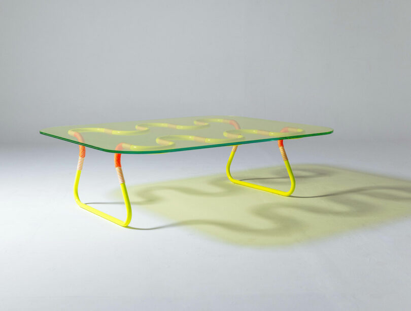 A rectangular glass coffee table by Laura Casañas Maya features a wavy yellow and orange metal frame, casting a patterned shadow on a plain white background.