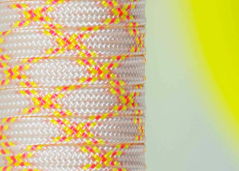 Close-up of a spool of braided rope designed by Laura Casañas Maya, featuring a white base with yellow and pink crisscross patterns, set against a blurred yellow and gray background.