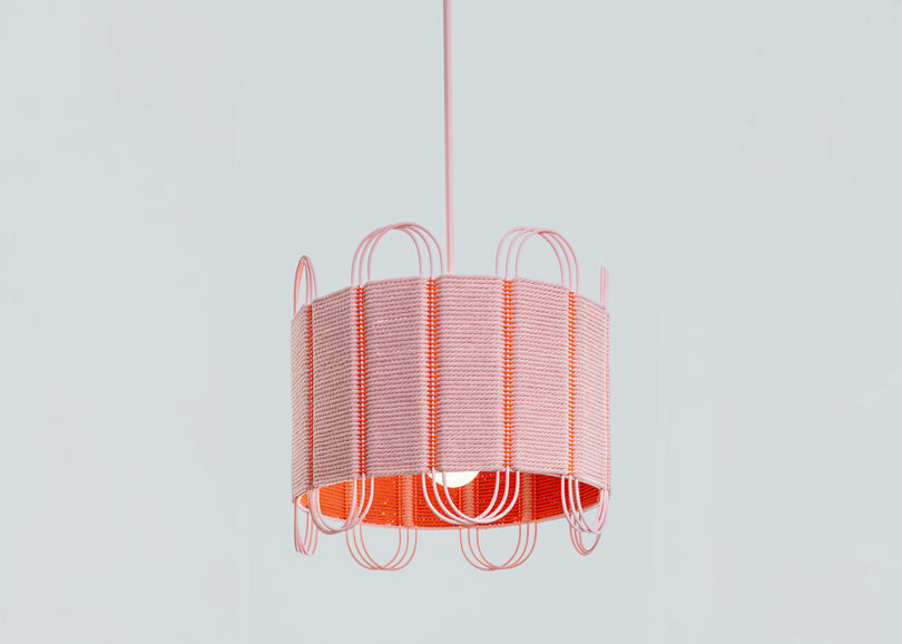 A pink and orange pendant light by Laura Casañas Maya features a woven cylindrical shade and decorative curved wire accents, hanging against a plain grey background.
