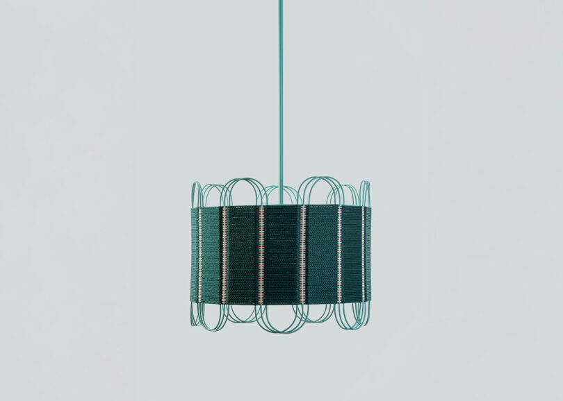 A modern pendant light by Laura Casañas Maya, featuring a cylindrical dark shade and an outer frame of teal wire loops, suspended from a matching teal cord against a light gray background.