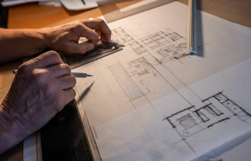 A person sketches architectural drawings on tracing paper using a ruler and mechanical pencil at a work desk.