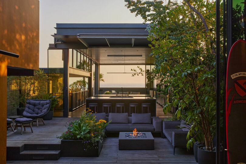 Contemporary outdoor patio with modern furniture, a fire pit, lush plants, and an open kitchen area facing the ocean at sunset.