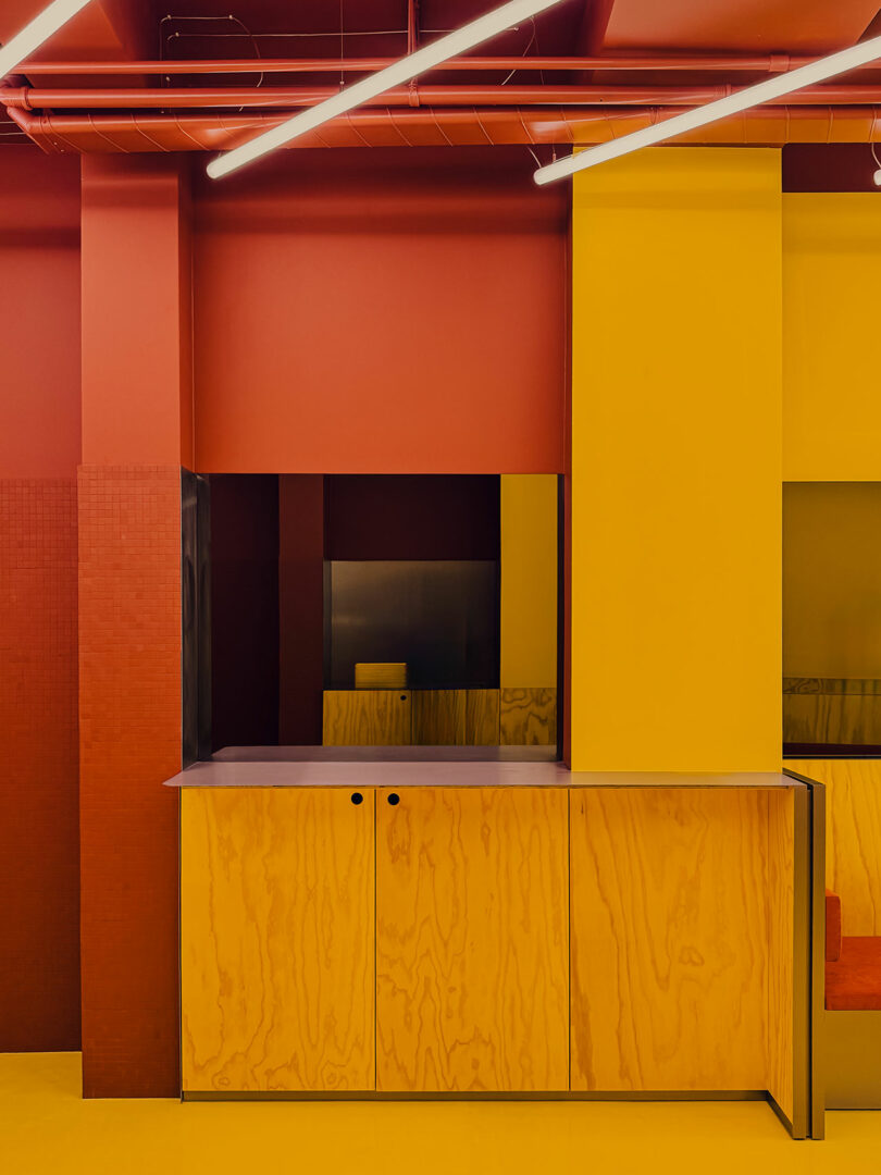 A minimalist interior with yellow walls and cabinetry, red ceiling pipes, and linear ceiling lights, featuring natural wood textures and geometric design elements.