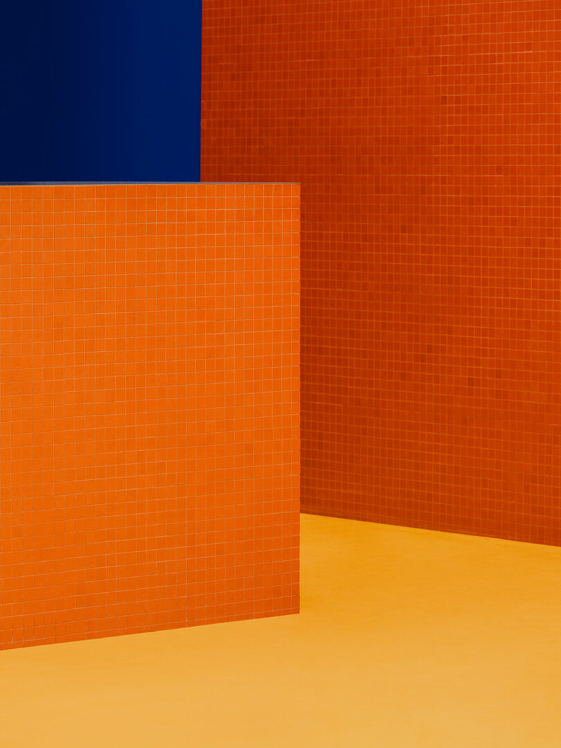 Photo of a room with orange tiled walls and floor, featuring a section of deep blue wall in the upper left corner. Geometric, minimalist design.