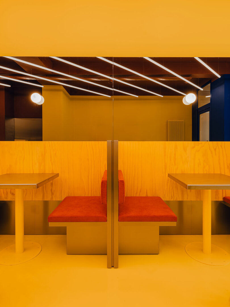 A modern seating booth with orange cushioned seats, yellow walls, and tables, set under linear ceiling lights and round wall lamps.
