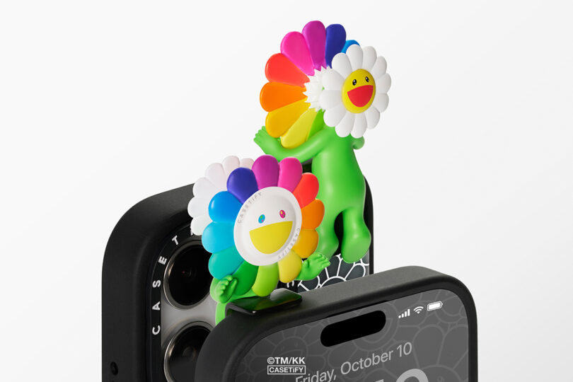 Two colorful, flower-shaped phone charms are attached to the top and camera area of a black phone case with a spiderweb design.