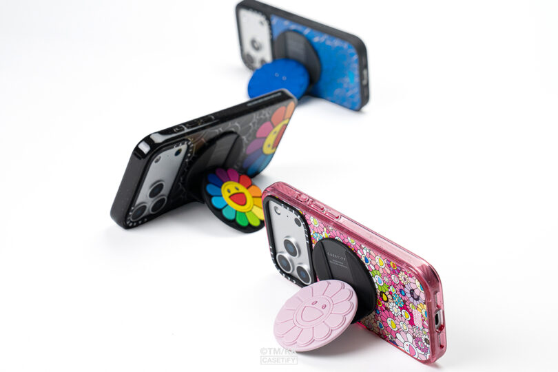 Three smartphones with floral-designed pop sockets are displayed upright on a white surface, each in different colored cases: black, blue, and pink.