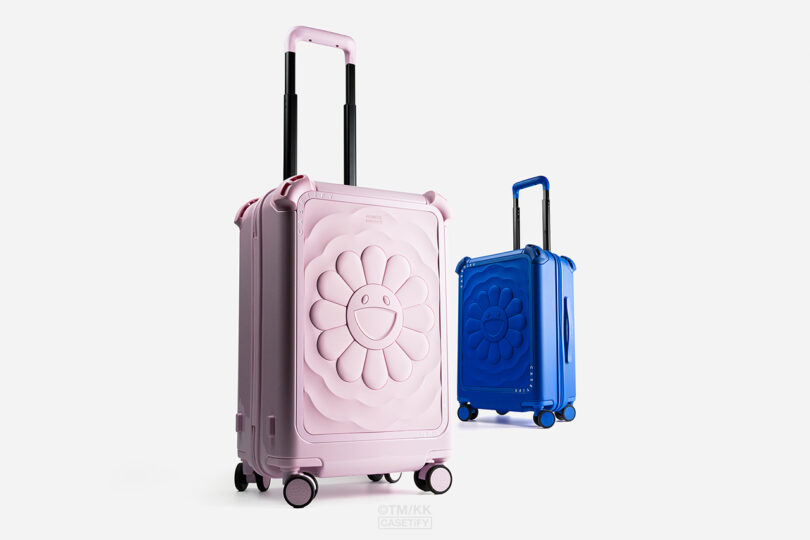 Two hard-shell suitcases, one pink and one blue, each with a raised flower design and smiley face on the front, stand upright with extended handles.