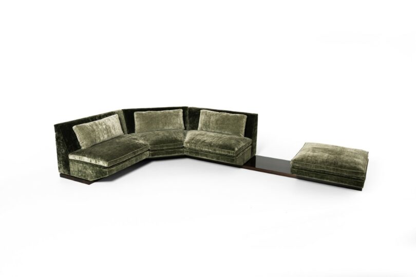 Green velvet sectional sofa with three seats, an attached wooden side table, and a matching ottoman, all set against a white background