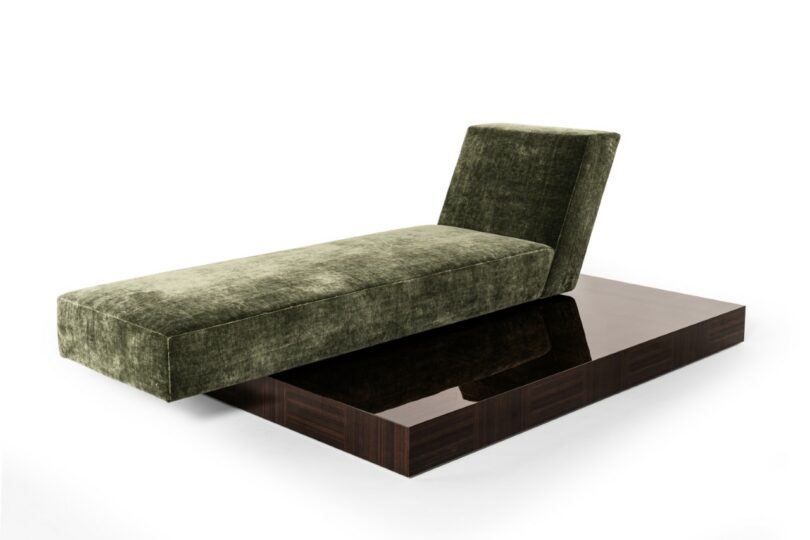 A modern chaise lounge with a green velvet cushion and angled backrest sits on a dark, glossy wooden platform against a white background