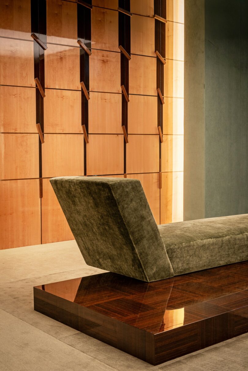 A modern lounge chair with a green upholstered seat sits on a polished wooden base in front of a geometric wood-paneled wall