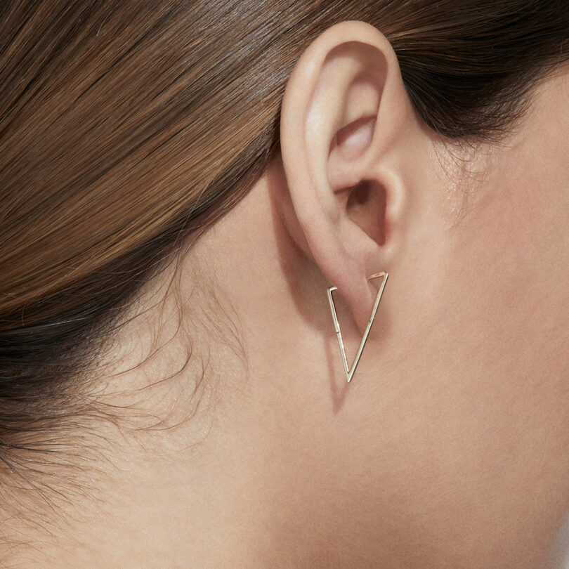 Close-up of a person’s ear wearing a minimalist, V-shaped gold earring. The person's hair is tucked behind the ear.