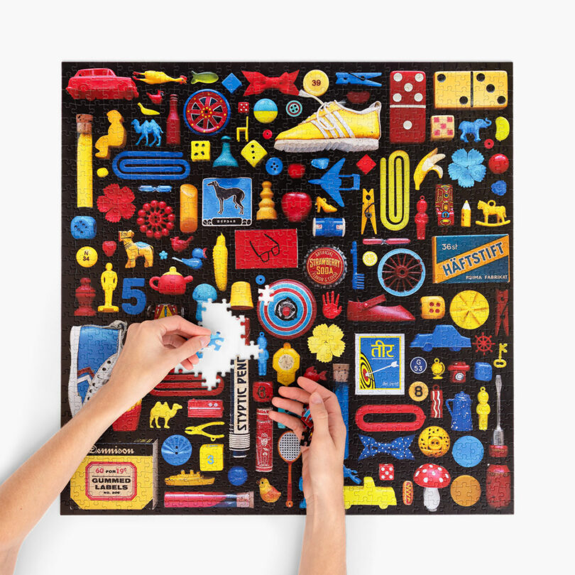 Two hands place a puzzle piece into a colorful jigsaw puzzle featuring assorted vintage objects in red, blue, and yellow, neatly arranged on a black background.