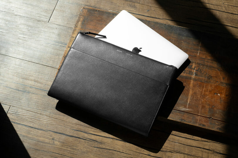 A closed black leather laptop sleeve partially covering an open silver MacBook on a wooden surface in sunlight.