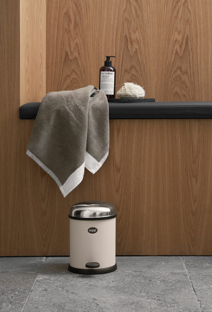 A beige pedal bin sits on a gray tiled floor below a black ledge holding a folded towel, a bottle of soap, and a natural scrub, against a wooden wall.