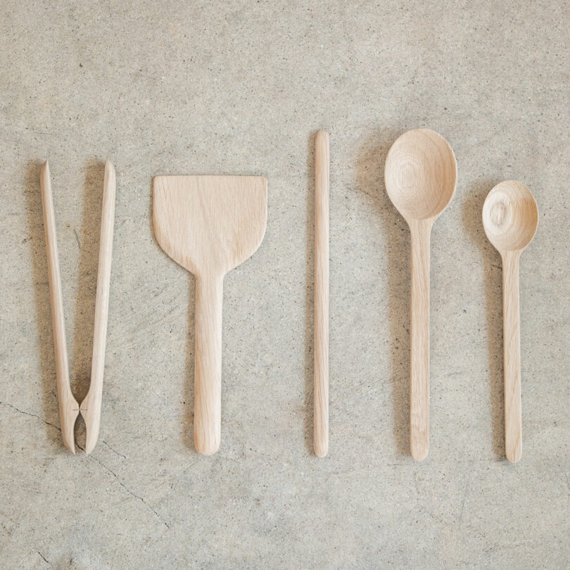 Five wooden kitchen utensils, perfect as gifts for cooks, including tongs, a flat spatula, a stick, a large spoon, and a small spoon, are arranged in a row on a light-colored textured surface.
