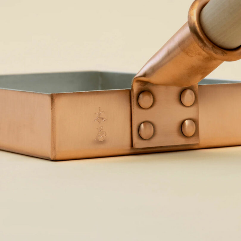 Close-up of a square copper pan with Japanese characters engraved on the side and a riveted handle attached, making it a unique addition to any kitchen and an ideal choice for gifts for cooks.