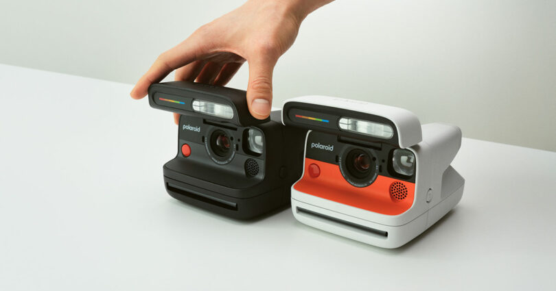 A hand touches the top of a black Polaroid instant camera next to a white Polaroid instant camera on a white surface, highlighting classic technology in a modern setting.