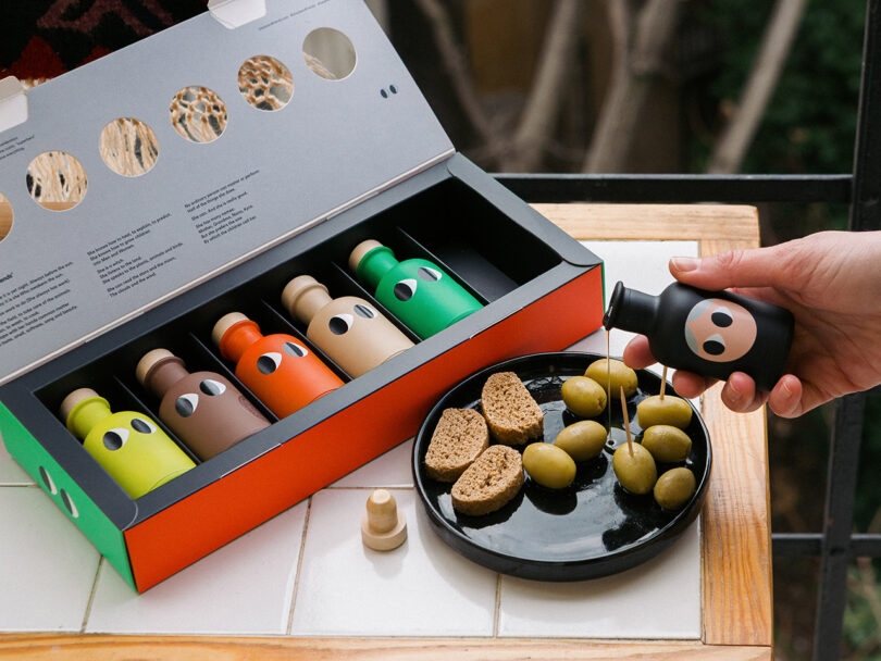 A hand pours oil from a small black bottle onto a plate with green olives and bread; an open box with five colorful bottles is on the table.