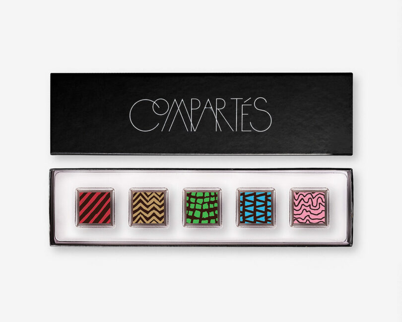 A black Compartés box with five square chocolates, each decorated with a distinct colorful geometric pattern, arranged in a row inside.