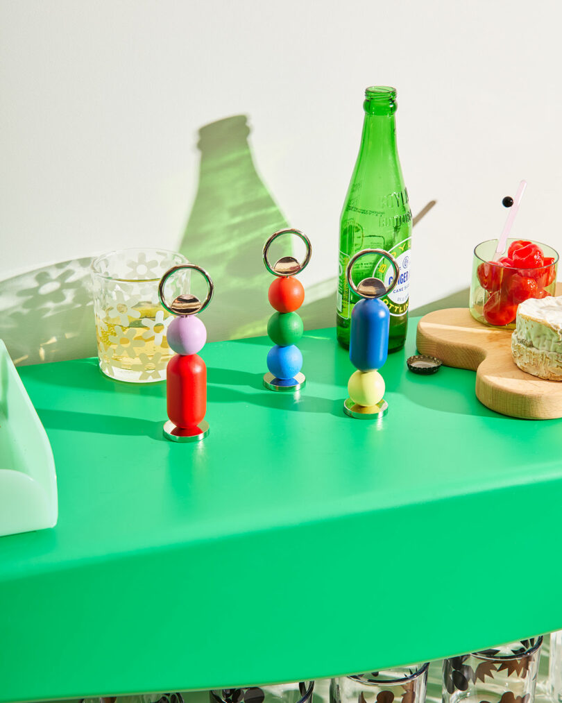 A green table with colorful bottle openers, a glass with yellow dots, a green bottle, a glass with skewered fruit, and a wooden tray holding cheese and bread.