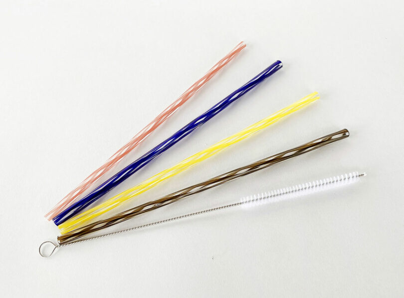 Three reusable straws in pink, blue, and yellow, one metal straw, and a cleaning brush are arranged on a white background.