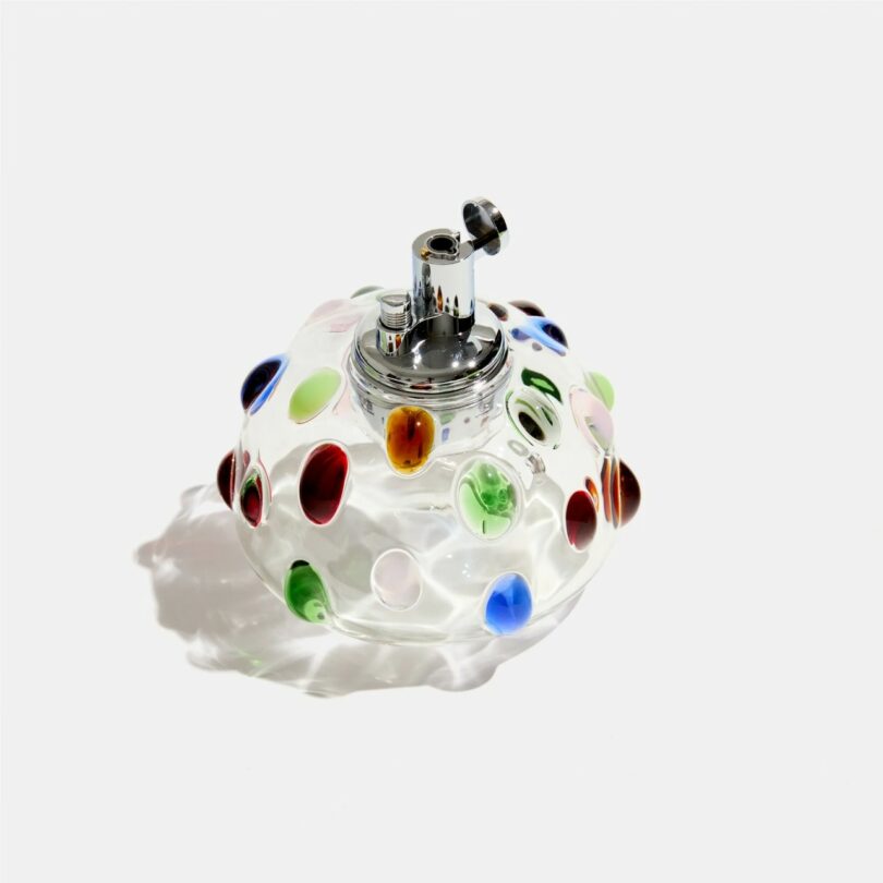 A clear glass table lighter with a wavy shape, decorated with colorful raised dots in red, blue, green, and yellow, on a plain white background