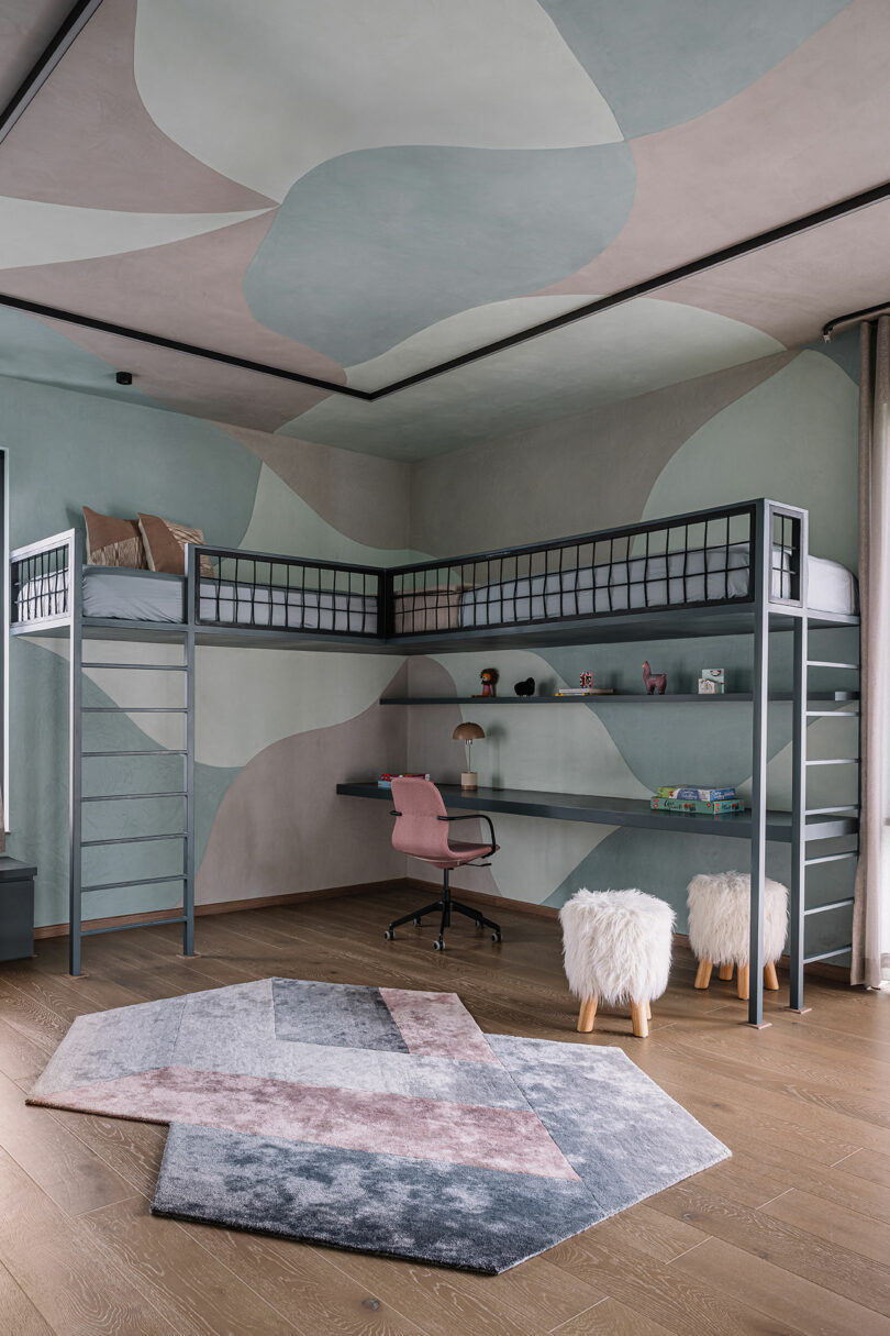 Modern bedroom with a loft bed, built-in desk, pink chair, two furry stools, geometric rug, shelves with decor, and abstract wall and ceiling patterns.