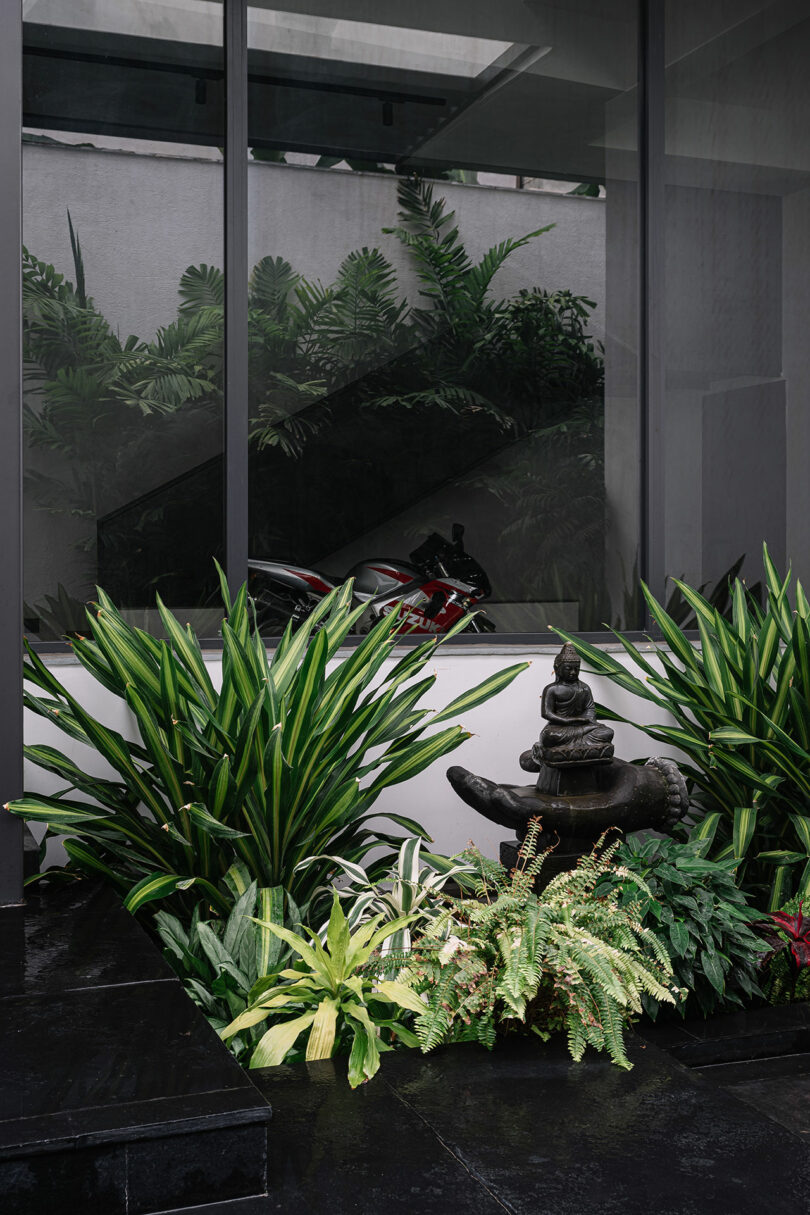 A modern outdoor area with lush green plants, a small statue on a pedestal, reflective windows, and a motorcycle visible in the background.